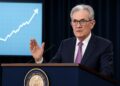 Jerome Powell gesticula ante podio de la Fed con gráfico económico ascendente de fondo.