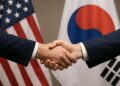Dos manos estrechándose con banderas de Estados Unidos y Corea del Sur desenfocadas al fondo.