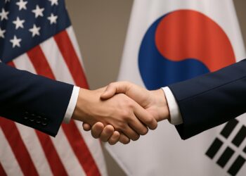 Dos manos estrechándose con banderas de Estados Unidos y Corea del Sur desenfocadas al fondo.