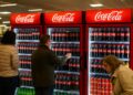 Clientes eligen bebidas frente a refrigeradores de Coca-Cola en un supermercado europeo iluminado.