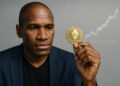 Arthur Hayes sostiene una moneda de Ethereum dorada con gesto serio sobre fondo gris.