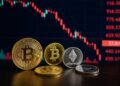 Varias monedas de Bitcoin y Ether en una mesa con un tablero digital al fondo mostrando caídas en rojo.