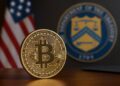 Moneda física de Bitcoin con bandera de EE. UU. y sello del Tesoro al fondo.