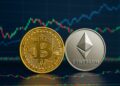 Monedas físicas de Bitcoin y Ethereum sobre mesa con pantallas mostrando gráficos financieros al fondo