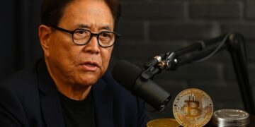 Robert Kiyosaki en un pódcast con micrófonos y monedas de Bitcoin, oro y plata en la mesa.