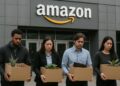 Cuatro trabajadores de Amazon con cajas y expresión triste salen de la sede con fachada gris industrial
