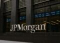 Edificio de JPMorgan en Nueva York con logo visible y pantallas de cotizaciones bursátiles al fondo