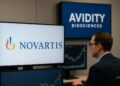 Hombre en oficina observa pantallas con logos de Novartis y Avidity Biosciences junto a gráficos financieros
