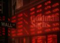 Pantallas rojas de cotizaciones en Wall Street con el logo difuminado de Goldman Sachs al fondo
