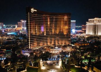 Torre curva del Wynn Las Vegas de noche con luces doradas cálidas, fuentes activas y huéspedes llegando por jardines, fondo del Strip visible