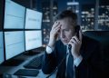 Un hombre estresado en traje en una oficina oscura por la noche, sosteniendo un teléfono y tocándose la frente. Detrás de él, varios monitores muestran los mensajes "NO DATA" y "DISCONNECTED".