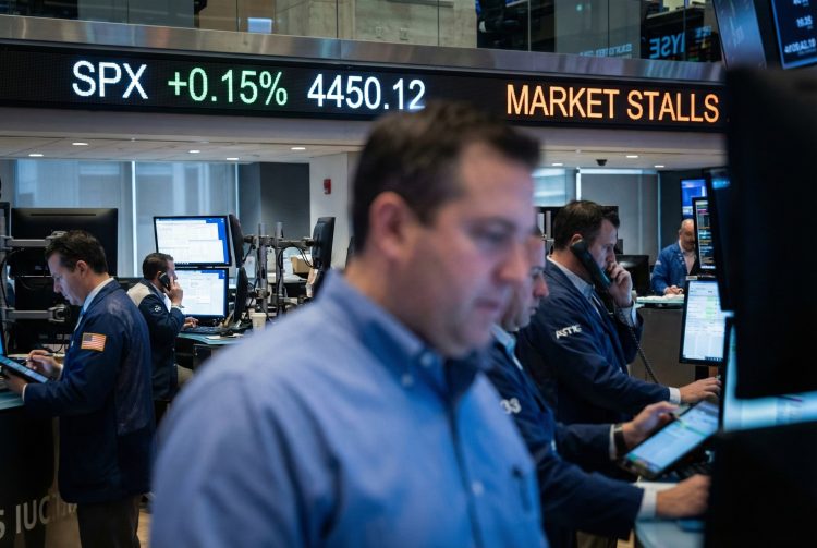 Interior de un piso de remates activo; una pantalla LED al fondo muestra "SPX +0.15%" y "MARKET STALLS" entre operadores de Bank of america trabajando.