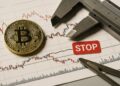 Moneda física de Bitcoin sobre gráfico bajista con relieve y etiqueta roja de alerta técnica.