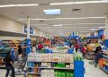 Interior amplio de un supermercado Walmart con familias comprando, empleados reponiendo estanterías y carritos en movimiento bajo iluminación LED brillante