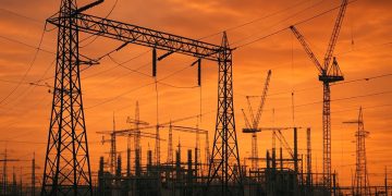 Vista panorámica de subestación eléctrica con grúas y nuevas líneas de transmisión en construcción bajo cielo naranja del ocaso, reflejando expansión de red bajo presión energética