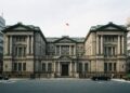 Fotografía arquitectónica frontal de la imponente fachada de piedra neoclásica del edificio del Banco de Japón en Tokio. La bandera nacional japonesa y la bandera institucional del banco ondean en el tejado, y una placa dorada sobre la entrada principal identifica claramente el edificio como NIPPON GINKO. Un peatón camina por la acera en primer plano.