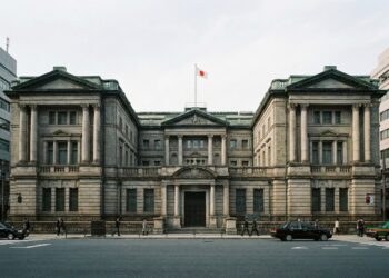 Fotografía arquitectónica frontal de la imponente fachada de piedra neoclásica del edificio del Banco de Japón en Tokio. La bandera nacional japonesa y la bandera institucional del banco ondean en el tejado, y una placa dorada sobre la entrada principal identifica claramente el edificio como NIPPON GINKO. Un peatón camina por la acera en primer plano.