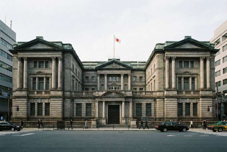 Fotografía arquitectónica frontal de la imponente fachada de piedra neoclásica del edificio del Banco de Japón en Tokio. La bandera nacional japonesa y la bandera institucional del banco ondean en el tejado, y una placa dorada sobre la entrada principal identifica claramente el edificio como NIPPON GINKO. Un peatón camina por la acera en primer plano.