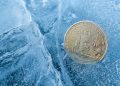 Primer plano de una moneda física de Bitcoin escarchada, parcialmente enterrada en hielo azul agrietado.