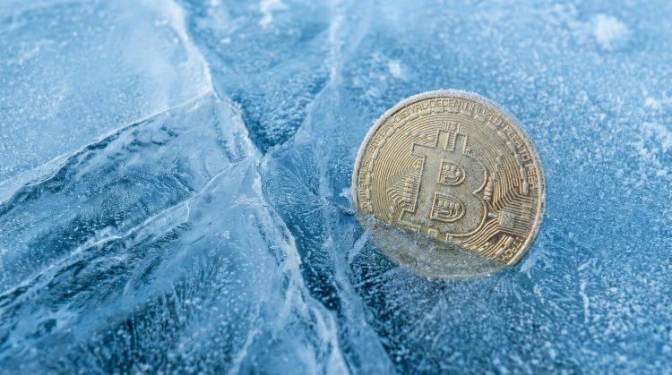 Primer plano de una moneda física de Bitcoin escarchada, parcialmente enterrada en hielo azul agrietado.