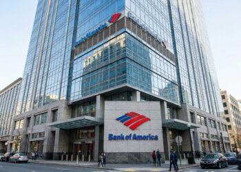 Fotografía corporativa exterior en un ángulo dinámico de tres cuartos de la torre sede de Bank of America en Charlotte. El logotipo de la bandera roja y azul y las letras 'Bank of America' son muy visibles sobre la señalización de piedra en la entrada a nivel de calle y en la parte superior de la fachada de vidrio reflectante bajo la luz del día.