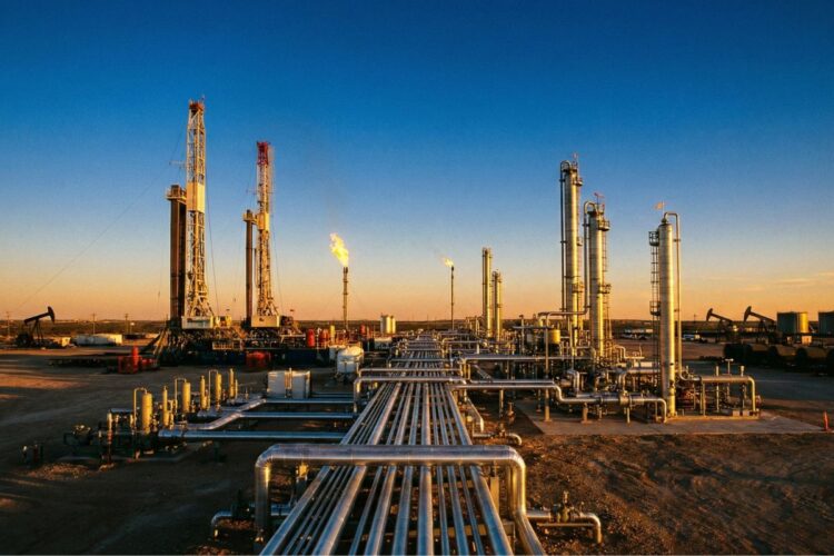 Vista panorámica al atardecer de una sofisticada instalación industrial de extracción de petróleo y gas en la Cuenca Pérmica. La luz dorada ilumina torres de perforación, chimeneas con llamas activas y una compleja red de tuberías metálicas en primer plano sobre un paisaje árido.