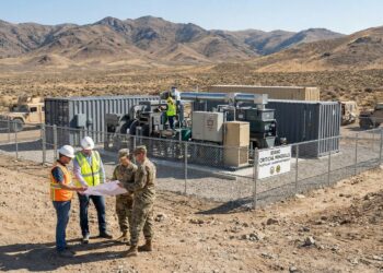 Cuatro hombres, dos ingenieros civiles con cascos y chalecos y dos soldados del Ejército de EE. UU. con uniforme de camuflaje, revisan planos frente a una planta piloto modular de procesamiento de minerales cercada en un paisaje desértico y montañoso. Un letrero identifica la instalación como "IDAHO CRITICAL MINERALS PILOT PLANT - US ARMY PARTNERSHIP". Vehículos militares están estacionados al fondo bajo la luz del día.