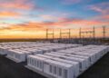 Fotografía panorámica al atardecer de una instalación masiva de Sistemas de Almacenamiento de Energía en Baterías (BESS) a escala de red. Cientos de contenedores blancos modulares de baterías (algunos con el logotipo visible de Tesla) están dispuestos en filas ordenadas, conectados a una infraestructura de subestación eléctrica de alta tensión con torres de transmisión en el fondo, bajo un cielo con tonos naranjas y rosados.