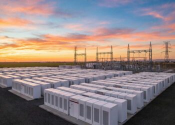 Fotografía panorámica al atardecer de una instalación masiva de Sistemas de Almacenamiento de Energía en Baterías (BESS) a escala de red. Cientos de contenedores blancos modulares de baterías (algunos con el logotipo visible de Tesla) están dispuestos en filas ordenadas, conectados a una infraestructura de subestación eléctrica de alta tensión con torres de transmisión en el fondo, bajo un cielo con tonos naranjas y rosados.