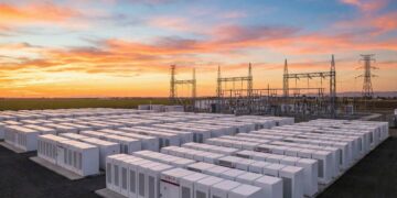 Fotografía panorámica al atardecer de una instalación masiva de Sistemas de Almacenamiento de Energía en Baterías (BESS) a escala de red. Cientos de contenedores blancos modulares de baterías (algunos con el logotipo visible de Tesla) están dispuestos en filas ordenadas, conectados a una infraestructura de subestación eléctrica de alta tensión con torres de transmisión en el fondo, bajo un cielo con tonos naranjas y rosados.