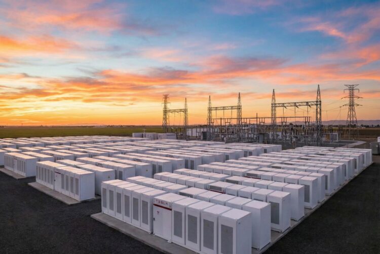 Fotografía panorámica al atardecer de una instalación masiva de Sistemas de Almacenamiento de Energía en Baterías (BESS) a escala de red. Cientos de contenedores blancos modulares de baterías (algunos con el logotipo visible de Tesla) están dispuestos en filas ordenadas, conectados a una infraestructura de subestación eléctrica de alta tensión con torres de transmisión en el fondo, bajo un cielo con tonos naranjas y rosados.