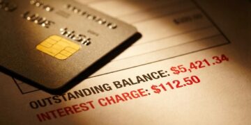 Fotografía de bodegón financiero con iluminación dramática y cálida. En primer plano, el chip dorado de una tarjeta de crédito descansa sobre un estado de cuenta impreso desenfocado. Un enfoque macro resalta números en tinta roja que indican saldos pendientes e intereses acumulados, visualizando la presión de la deuda sobre la economía doméstica.