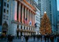 Fotografía documental al atardecer que muestra la fachada neoclásica de la Bolsa de Nueva York (NYSE) en Broad Street, decorada con guirnaldas y luces festivas. Un gran árbol de Navidad iluminado se alza a la derecha del edificio, y las banderas de EE. UU. y de la NYSE ondean en las columnas mientras peatones caminan por la calle en un ambiente invernal.