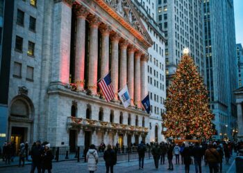 Fotografía documental al atardecer que muestra la fachada neoclásica de la Bolsa de Nueva York (NYSE) en Broad Street, decorada con guirnaldas y luces festivas. Un gran árbol de Navidad iluminado se alza a la derecha del edificio, y las banderas de EE. UU. y de la NYSE ondean en las columnas mientras peatones caminan por la calle en un ambiente invernal.