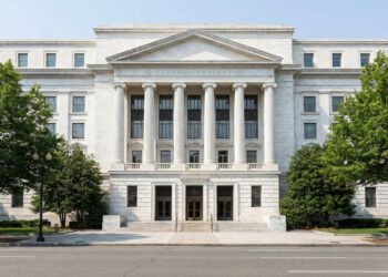 Fotografía frontal y simétrica del edificio Marriner S. Eccles de la Reserva Federal en Washington D.C., mostrando su fachada de mármol blanco, columnas clásicas y el grabado "FEDERAL RESERVE" bajo luz diurna, flanqueado por árboles verdes.
