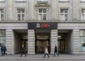 Fotografía documental a nivel de calle de la fachada de piedra clásica de una sede principal del banco UBS (probablemente en Zúrich), con el logotipo rojo y las llaves cruzadas visible sobre la entrada. Varios peatones caminan por la acera frente al edificio bajo luz natural, transmitiendo solidez institucional.