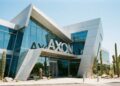 Vista frontal exterior a nivel de calle de la sede corporativa de Axon Enterprise en Scottsdale, Arizona. La imagen muestra una arquitectura moderna y angular con fachada de paneles metálicos plateados y grandes ventanales de vidrio. El logotipo 'AXON' en letras grandes y plateadas es prominente sobre la entrada principal, todo bajo luz diurna brillante y rodeado de paisaje desértico con cactus saguaro.