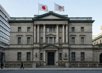 Fotografía arquitectónica frontal de la imponente fachada de piedra neoclásica del edificio del Banco de Japón en Tokio. La bandera nacional japonesa y la bandera institucional del banco ondean en el tejado, y una placa dorada sobre la entrada principal identifica claramente el edificio como NIPPON GINKO. Un peatón camina por la acera en primer plano.