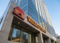 Fotografía en ángulo contrapicado de la fachada de una torre corporativa de Wells Fargo, mostrando el prominente logotipo rojo y dorado del carruaje y el nombre del banco sobre una estructura de granito y vidrio bajo luz diurna.