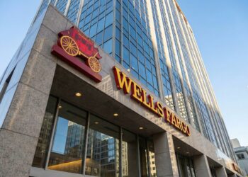 Fotografía en ángulo contrapicado de la fachada de una torre corporativa de Wells Fargo, mostrando el prominente logotipo rojo y dorado del carruaje y el nombre del banco sobre una estructura de granito y vidrio bajo luz diurna.