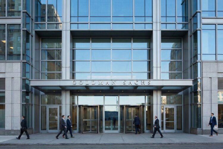 Vista frontal a nivel de calle de la entrada principal de la sede corporativa de Goldman Sachs en 200 West Street, Nueva York. La imagen captura la moderna fachada de vidrio y acero bajo luz diurna, con el nombre de la firma visible de forma sobria y profesionales financieros ingresando al edificio, simbolizando la solidez y actividad del banco de inversión.