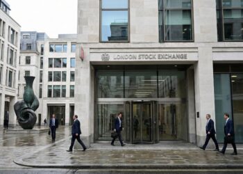 Fotografía documental exterior de la entrada principal de la London Stock Exchange (LSE) en Paternoster Square, Londres. Se observa la fachada de piedra y cristal con el nombre de la institución, varios profesionales con traje caminando por la plaza y la icónica escultura de bronce a la izquierda bajo un día nublado.