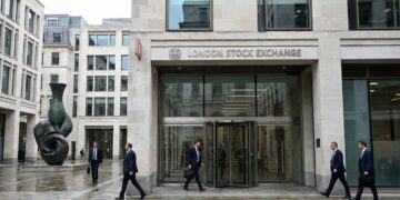 Fotografía documental exterior de la entrada principal de la London Stock Exchange (LSE) en Paternoster Square, Londres. Se observa la fachada de piedra y cristal con el nombre de la institución, varios profesionales con traje caminando por la plaza y la icónica escultura de bronce a la izquierda bajo un día nublado.