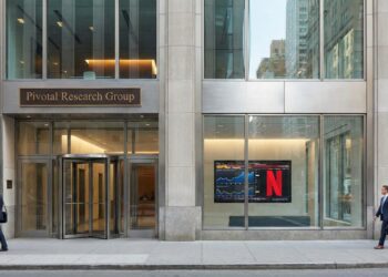 Fachada a nivel de calle de las oficinas de Pivotal Research Group. Un letrero de bronce sobre la entrada dice "Pivotal Research Group". Una pantalla en la ventana adyacente muestra gráficos financieros y el logotipo rojo "N" de Netflix. Peatones con trajes de negocios caminan por la acera frente al edificio de piedra y vidrio.