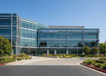 Fotografía exterior en un día soleado del moderno edificio de oficinas de Ross Stores. La fachada, construida principalmente de vidrio y paneles grises, muestra el logotipo azul "ROSS STORES" en la parte superior izquierda. La entrada principal, con el número 5130 visible, está rodeada de jardines bien cuidados, arbustos verdes y árboles bajo un cielo azul despejado. Una rotonda pavimentada conduce a la entrada del edificio.
