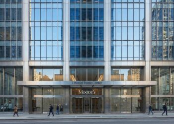 Vista frontal a nivel de calle de la entrada moderna de cristal y acero del edificio 7 World Trade Center. Los letreros "7 World Trade Center" y el logotipo "MOODY'S" son visibles sobre las puertas giratorias mientras peatones caminan por la acera.