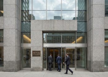 Fachada de granito y vidrio de Wolfe Research. Tres hombres de traje entran por la puerta giratoria.