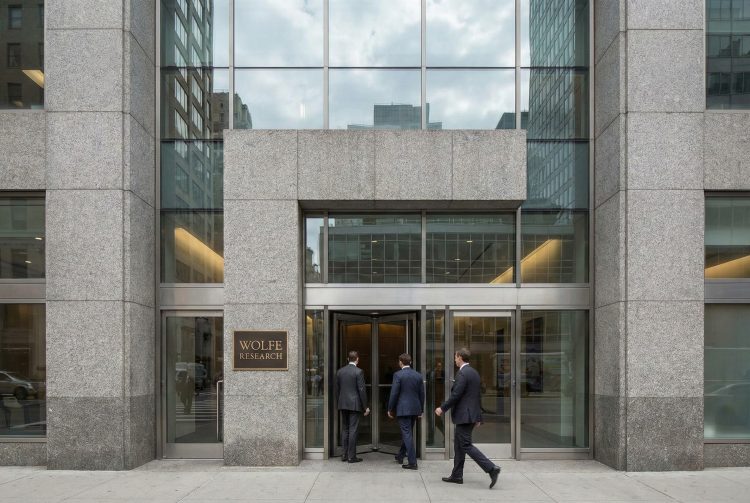 Fachada de granito y vidrio de Wolfe Research. Tres hombres de traje entran por la puerta giratoria.
