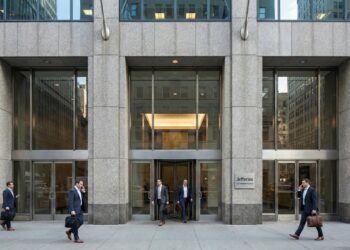 Vista frontal a nivel de calle de la entrada principal de la sede corporativa de Jefferies Financial Group en 520 Madison Avenue, Nueva York. La imagen muestra una fachada sólida de granito y vidrio bajo luz natural, con profesionales financieros entrando y saliendo del edificio, transmitiendo actividad institucional y autoridad en el mercado.