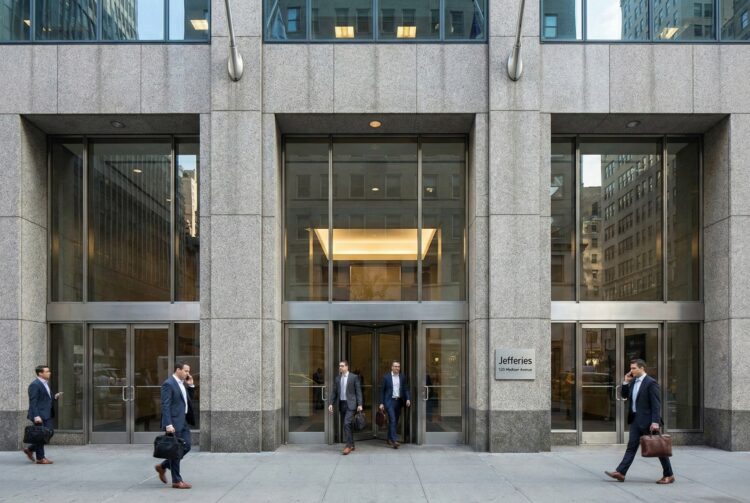 Vista frontal a nivel de calle de la entrada principal de la sede corporativa de Jefferies Financial Group en 520 Madison Avenue, Nueva York. La imagen muestra una fachada sólida de granito y vidrio bajo luz natural, con profesionales financieros entrando y saliendo del edificio, transmitiendo actividad institucional y autoridad en el mercado.